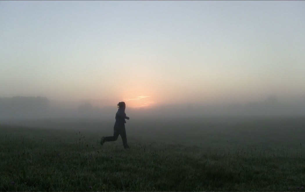 A person is jogging across a grassy field early in the morning. The landscape is covered in thick fog, blurring the horizon line.  We can see the red glow from a sunrise appearing amongst the misty background. The person running appears slightly mid air, in between steps. The sunrise makes them out like a silhouette on the land. The grass is covered in dew or frost.